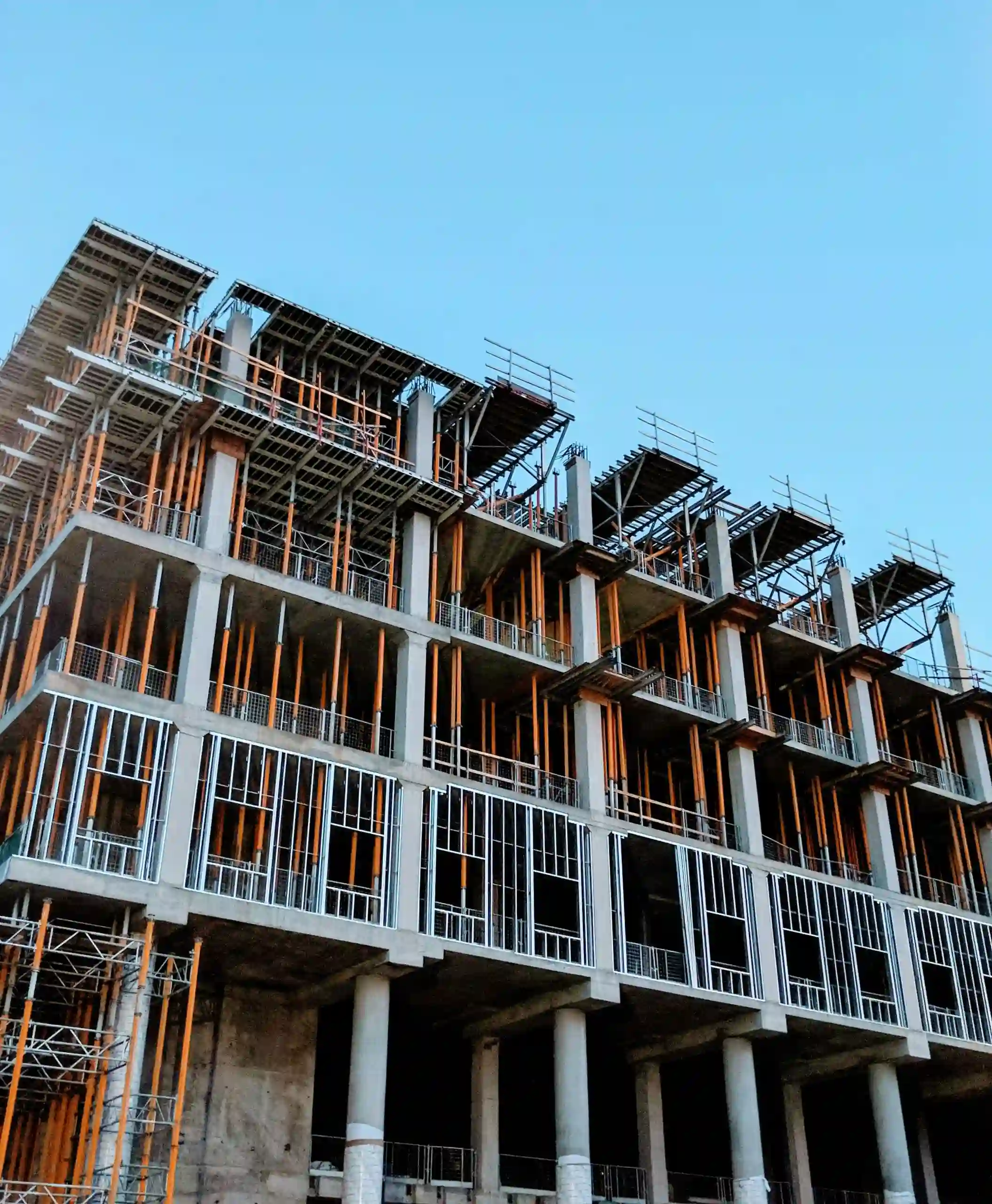 A multi-story building under construction with a concrete frame, supported by numerous orange scaffolding poles and temporary metal structures against a clear blue sky