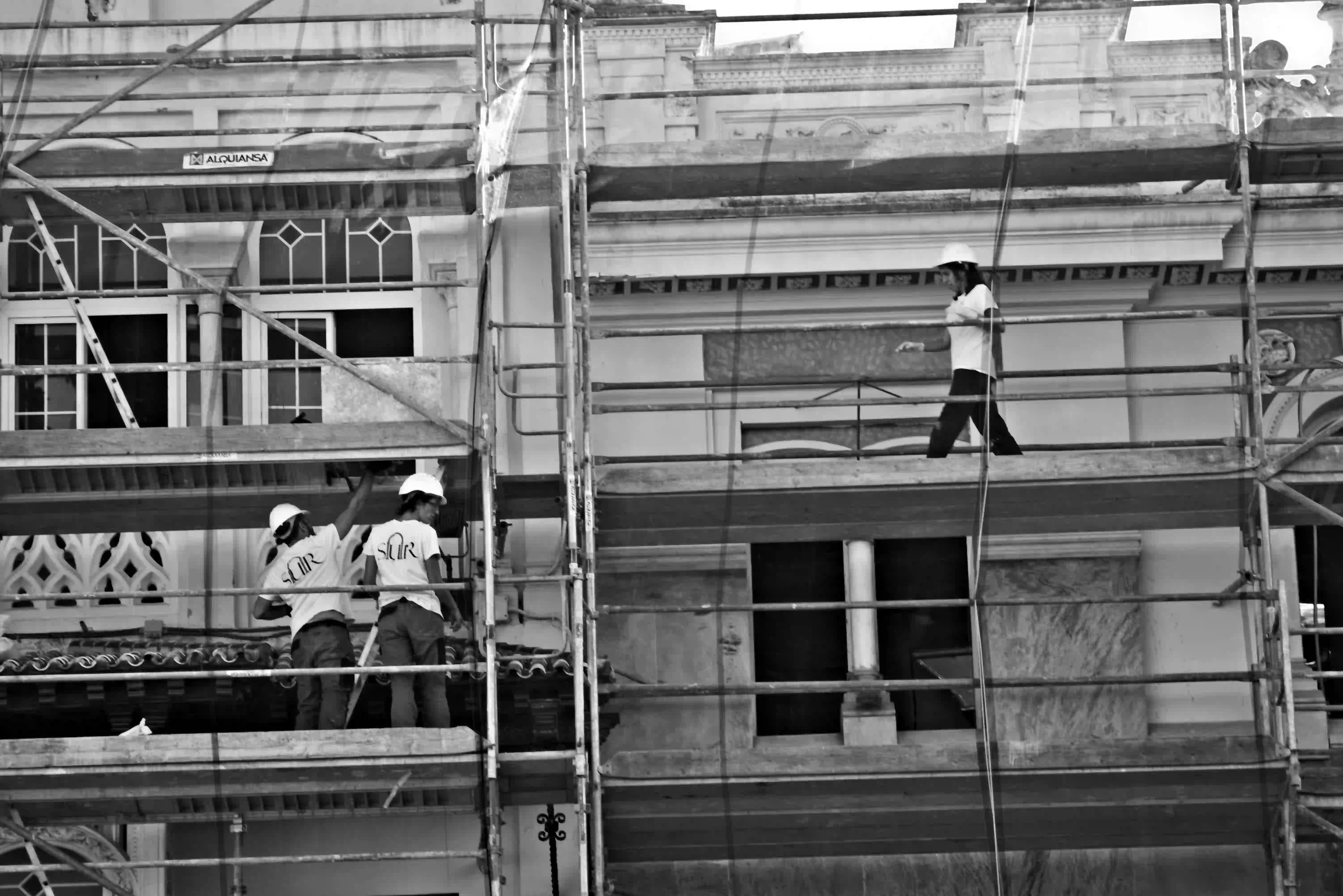 In this black-and-white photo, three construction workers in hard hats and white t-shirts are working on metal scaffolding in front of a classical building with ornate architectural details