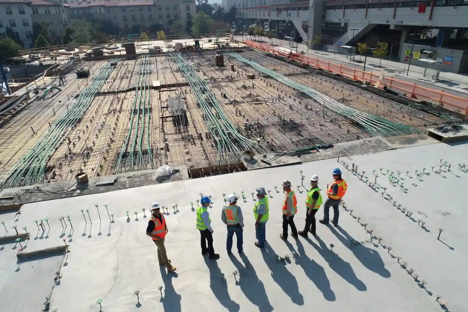 An aerial, high-angle view shows a group of construction workers in hard hats and safety vests standing on a finished concrete slab, looking over a large area of exposed rebar and green conduits laid out for the next phase of a building project