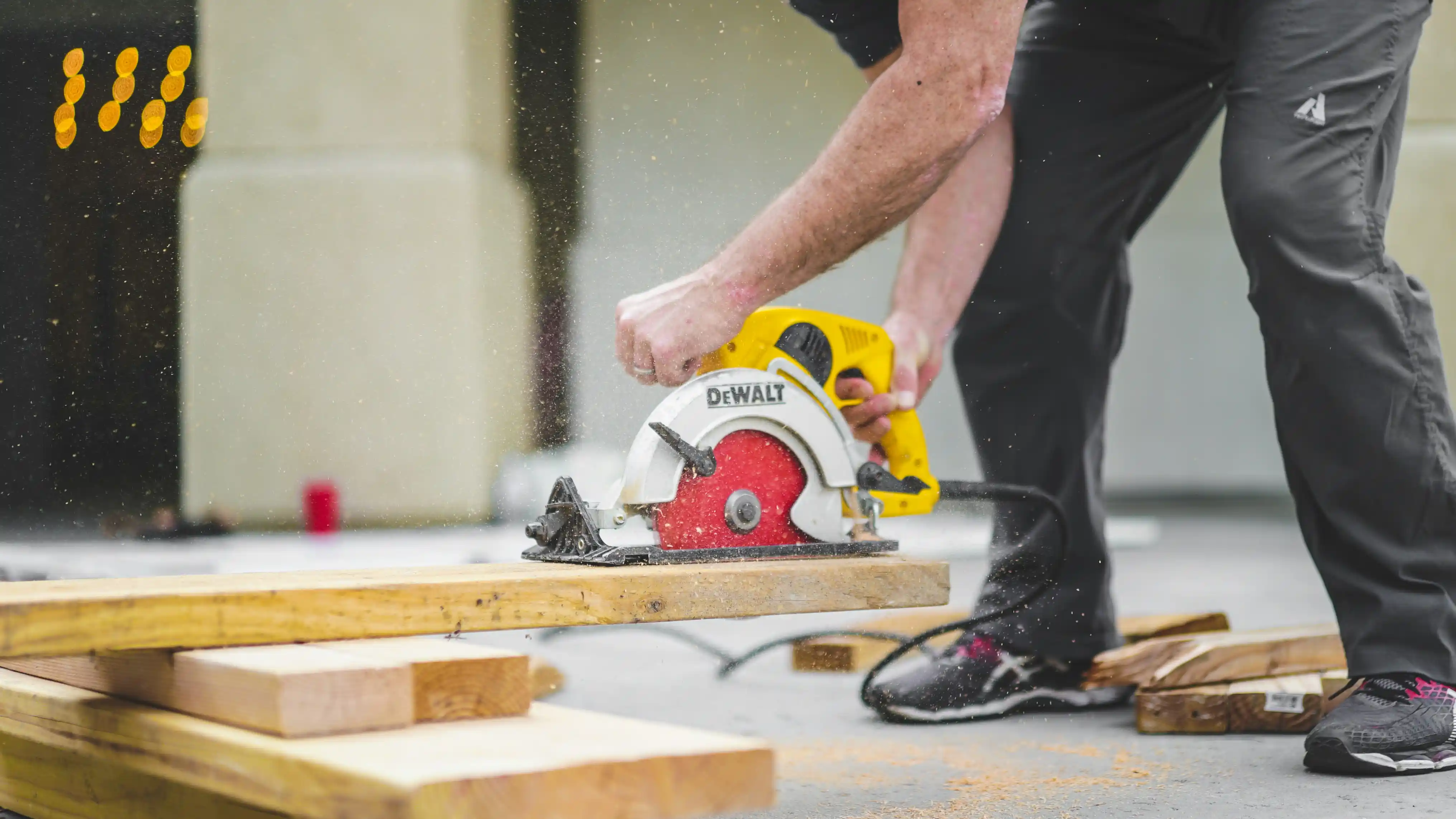 A worker's hands operate a yellow and silver DeWalt circular saw to cut through a thick wooden plank, sending sawdust into the air