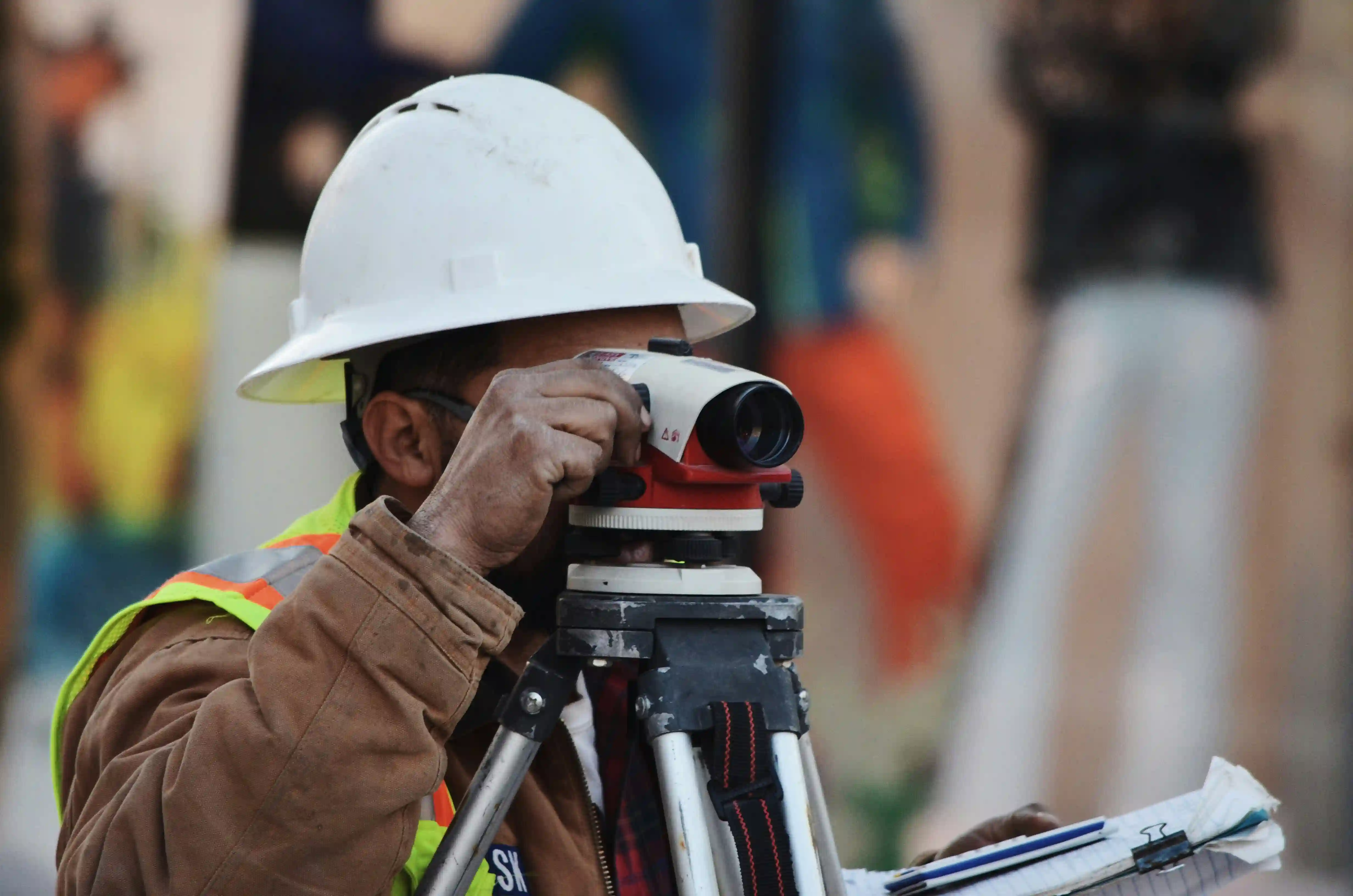 A close-up of a construction worker wearing a white hard hat and a brown jacket, looking through a professional surveyor's level mounted on a tripod while holding a notebook