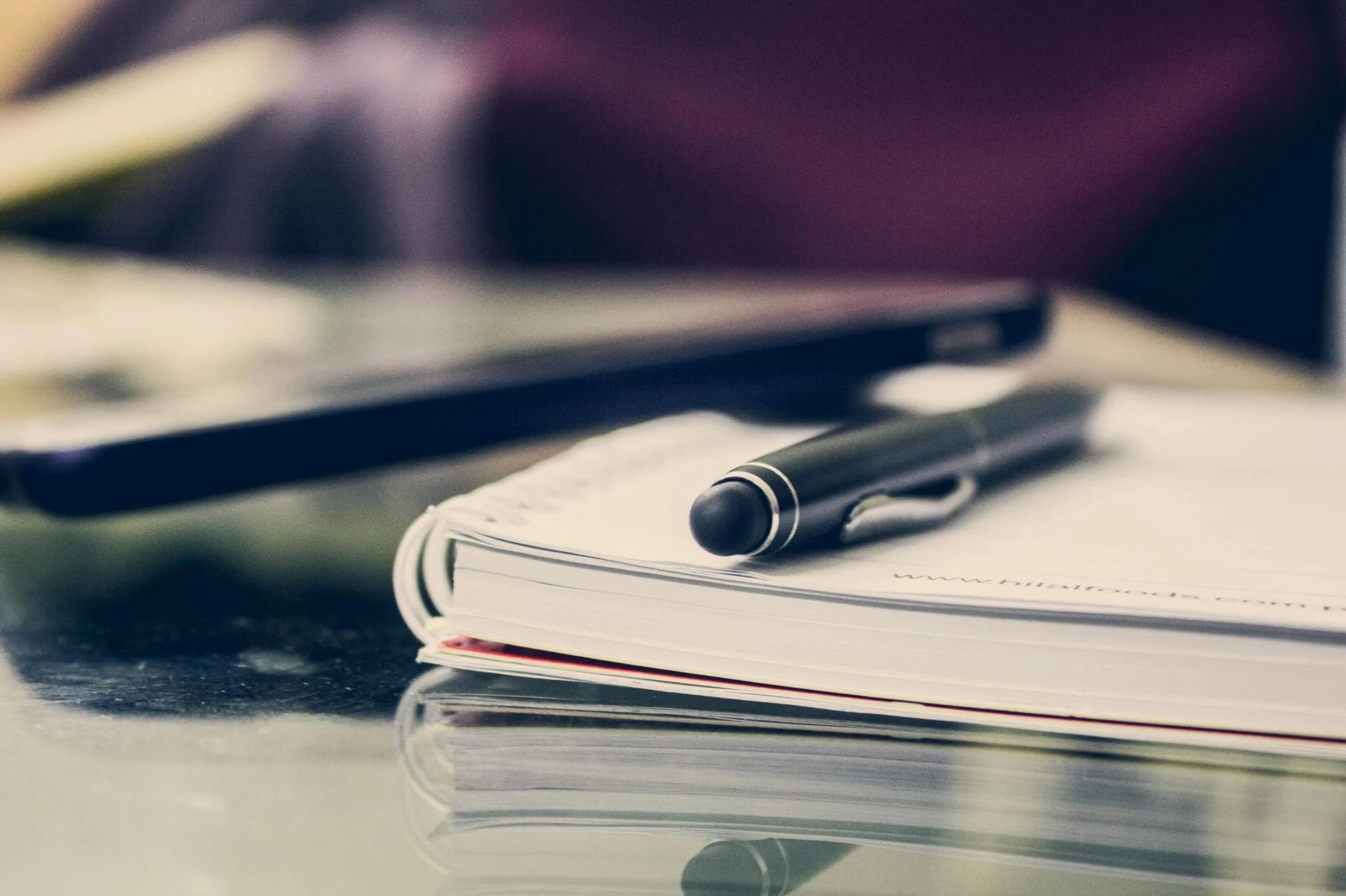 A close-up, shallow-depth-of-field shot of a black pen resting on top of a white spiral-bound notebook on a reflective glass surface