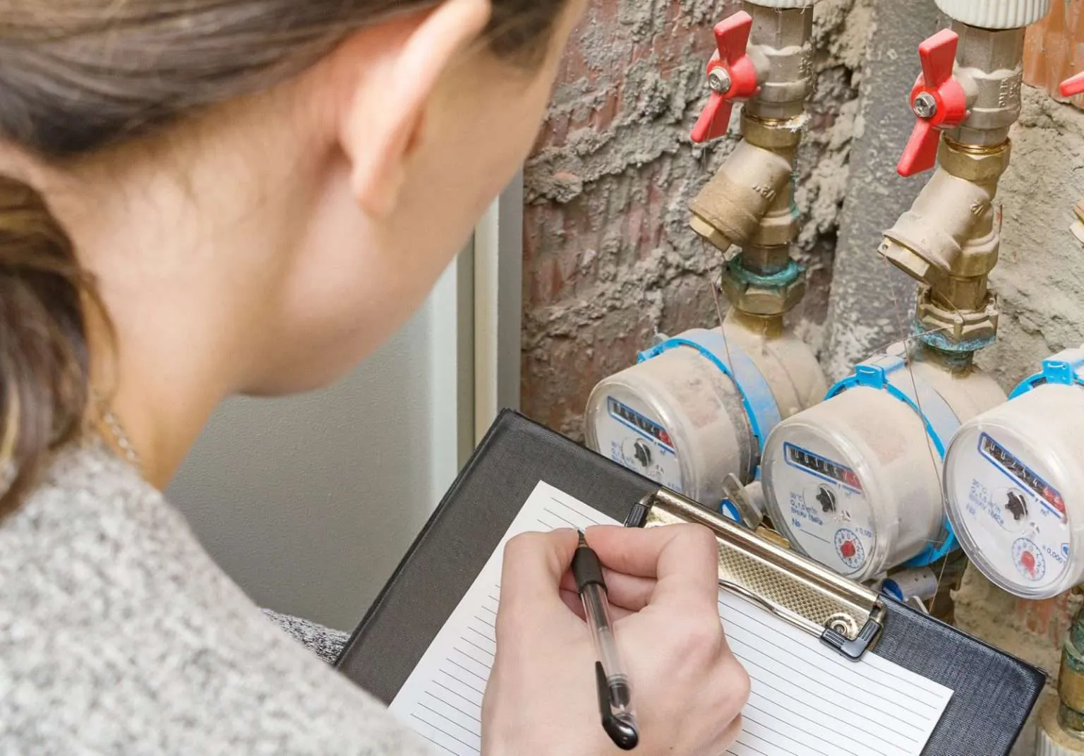 A woman seen from behind with her hair in a ponytail holds a black clipboard and writes on a piece of paper with a pen while looking at three water meters installed on a brick wall
