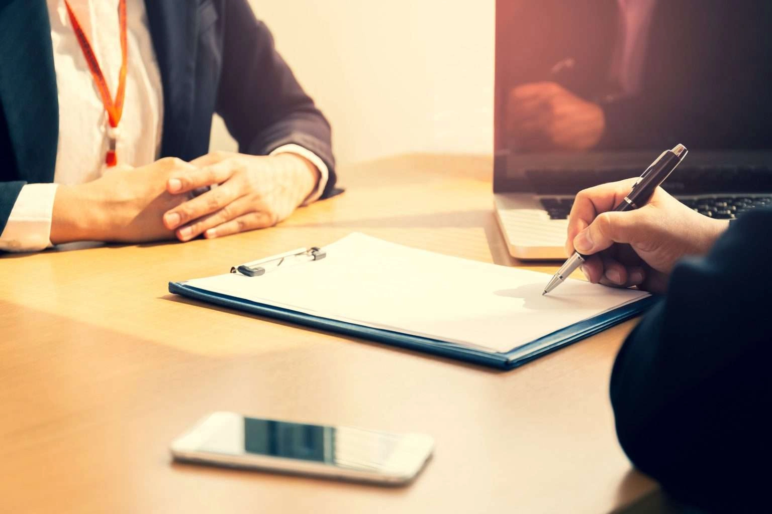 A person in professional attire sits at a desk across from another person whose hands are clasped. The foreground shows a hand holding a pen over a blank document on a blue clipboard, with a smartphone and a laptop also visible on the wooden table