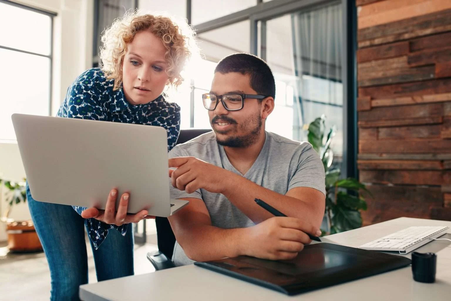 A woman with blonde curly hair stands behind a man with glasses and a beard, both looking intently at a laptop she is holding. The man is sitting at a desk with a digital drawing tablet and a stylus in his hand