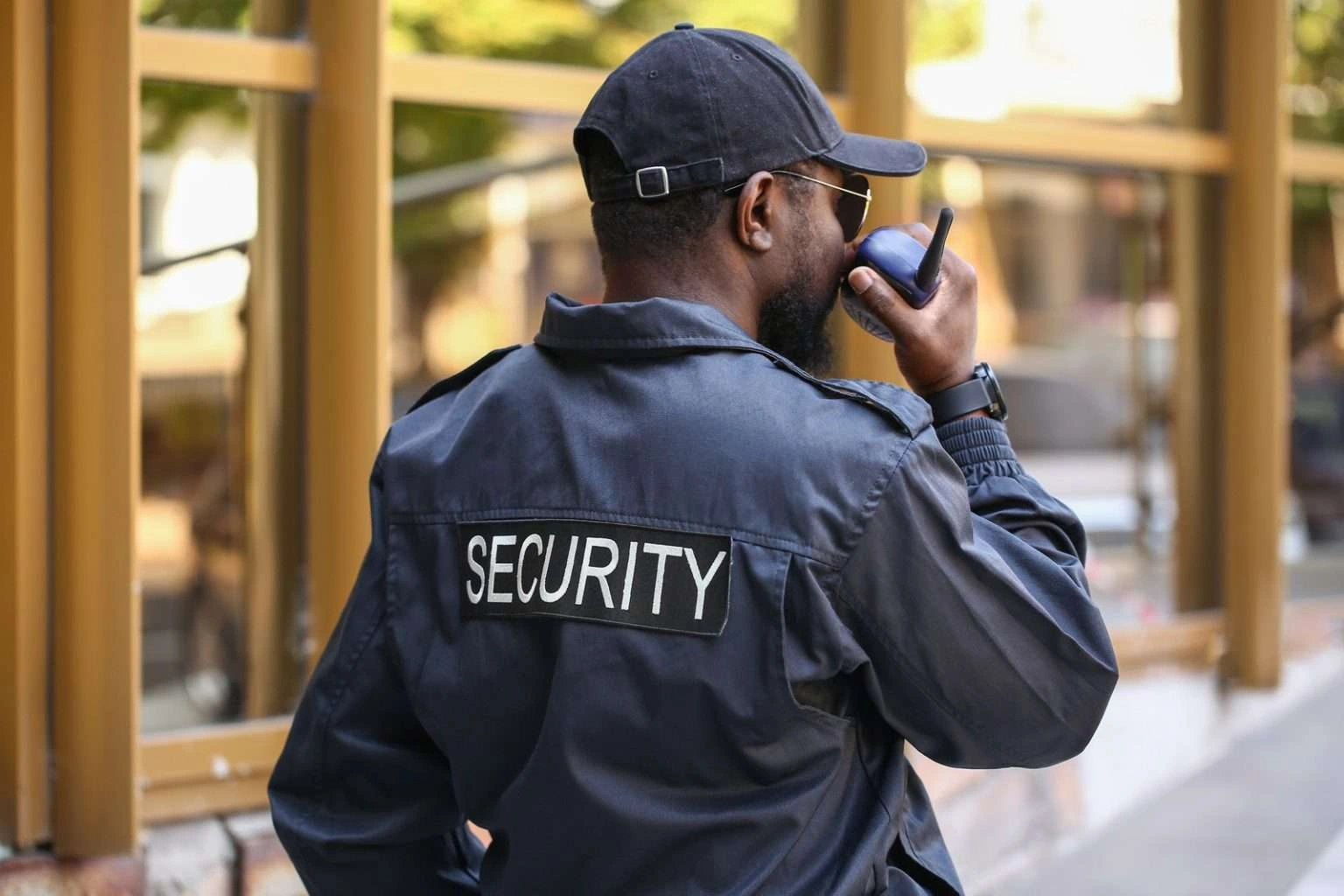 A man seen from behind wearing a navy blue security jacket with the word "SECURITY" in large white letters on the back. He is also wearing a black baseball cap and sunglasses while talking into a handheld radio