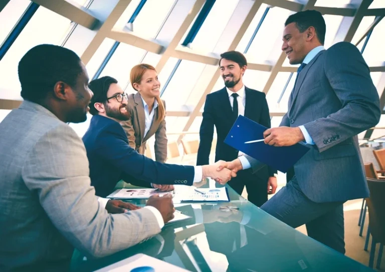 A group of business professionals in a modern office with large windows, where two men are shaking hands over a glass conference table while their colleagues look on and smile