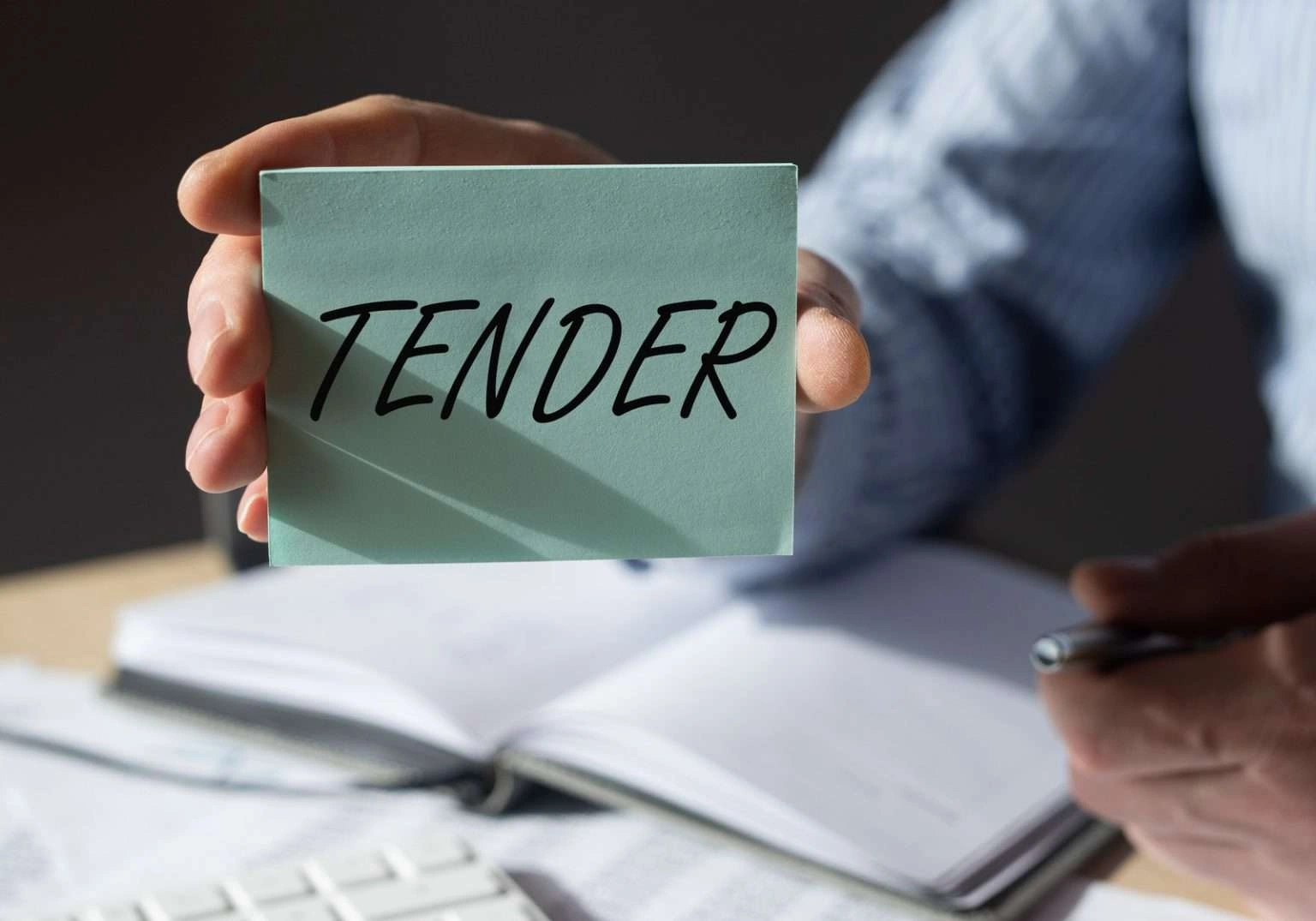 A person's hand holds a light green card with the word "TENDER" written on it in black ink. In the blurred background, a desk with an open notebook and a pen is visible