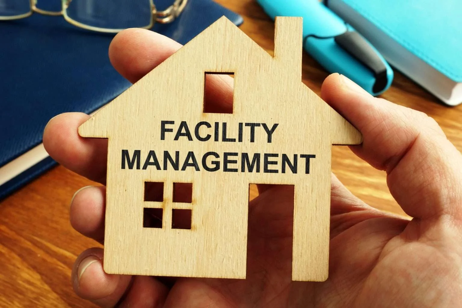 A person's hand holds a small wooden cutout in the shape of a house, which has the words "FACILITY MANAGEMENT" printed in black capital letters across the front