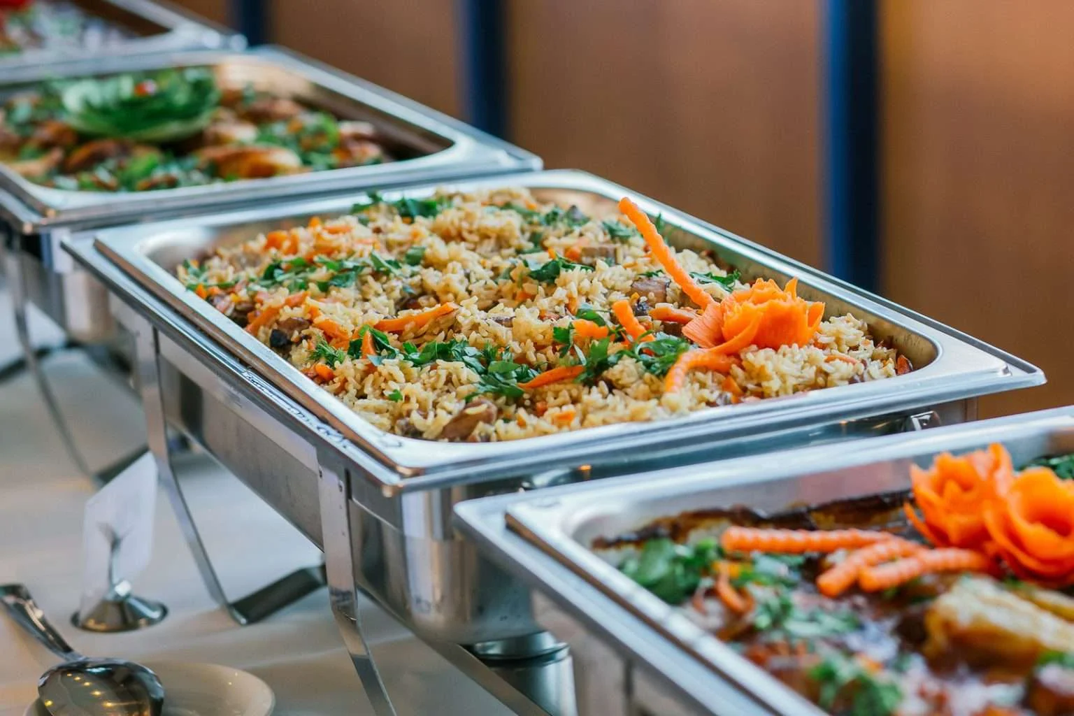 A row of silver catering chafing dishes filled with food, with the central dish containing a rice pilaf garnished with fresh herbs and decorative carrot carvings