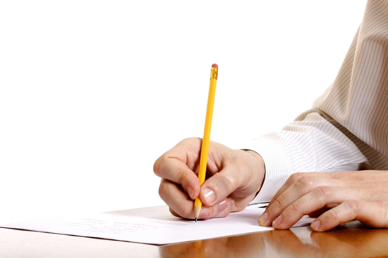 A close-up shot of a person's hand in a striped shirt, holding a yellow pencil and writing on a white sheet of paper on a wooden desk.