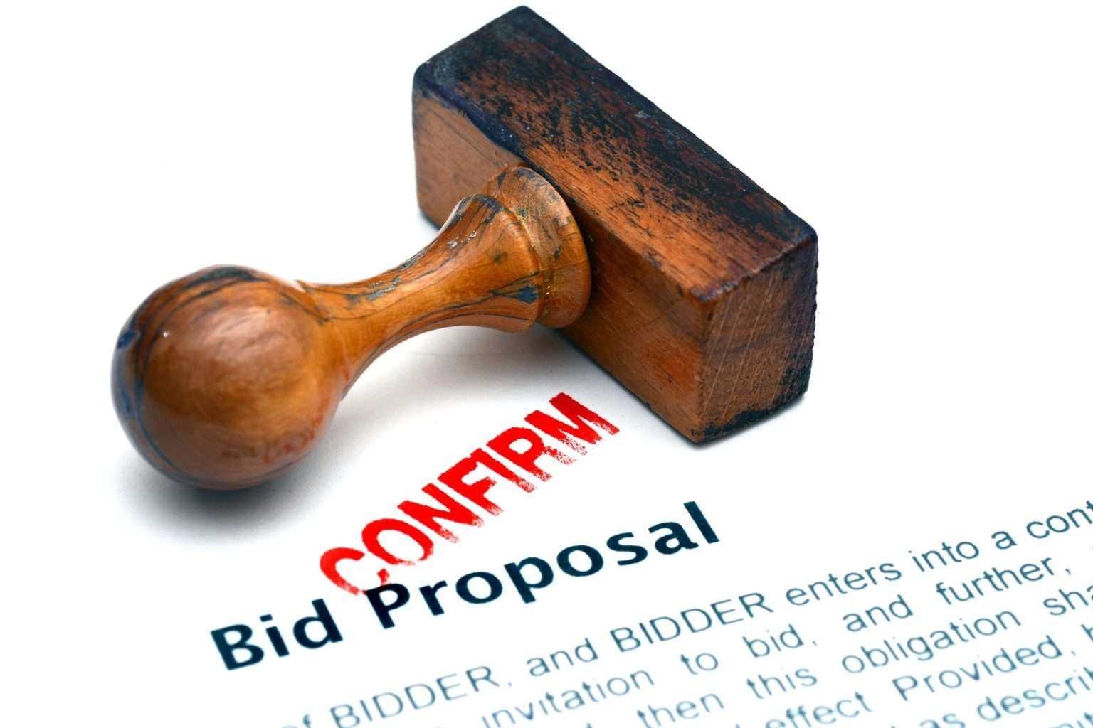 A wooden rubber stamp rests on a document titled "Bid Proposal," with a fresh red "CONFIRM" stamp mark visible on the white paper.