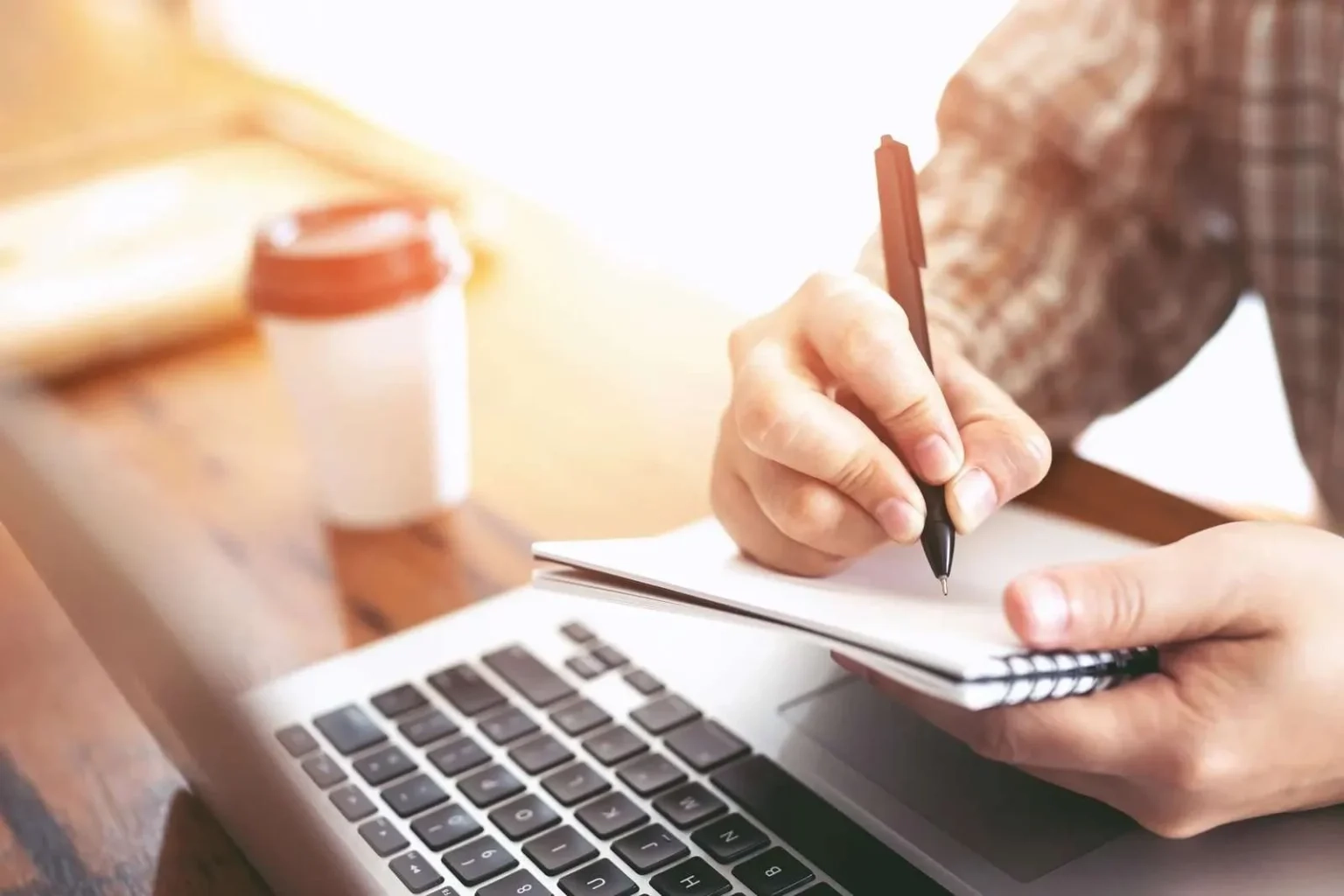 A close-up shot of a person's hand as they write in a small spiral notebook with a black pen, while sitting in front of an open laptop with a coffee cup in the background.