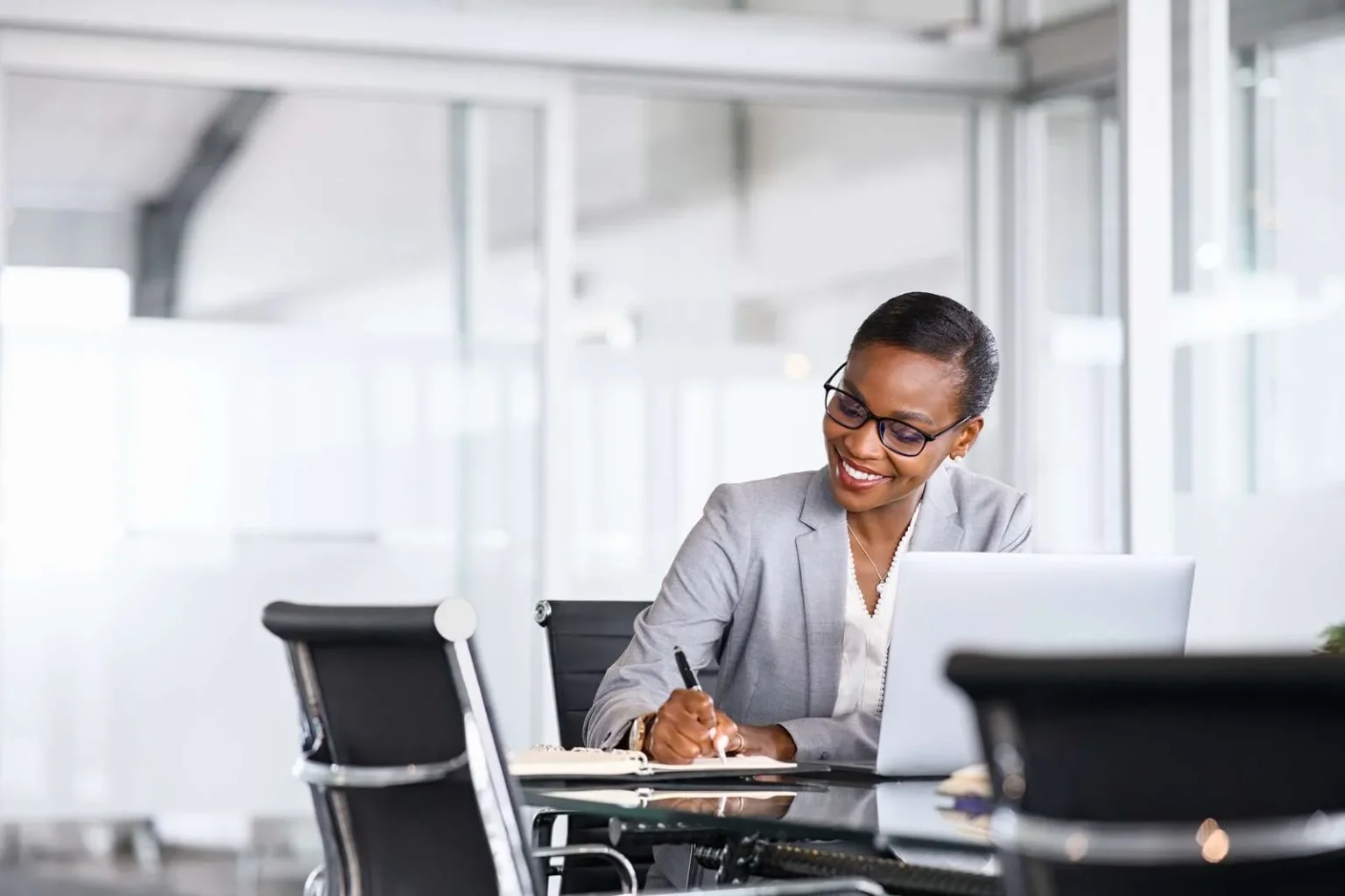 A smiling woman in a grey business suit and glasses sits at a glass desk in a bright, modern office, writing in a notebook while working on a laptop.
