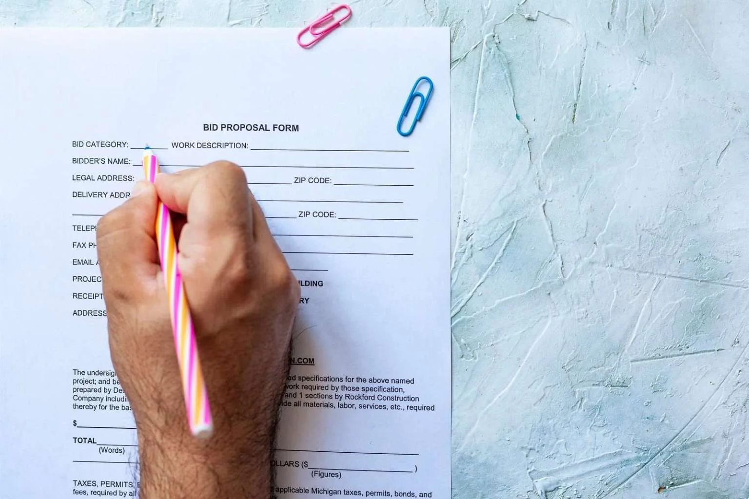 A top-down view of a hand using a pink and yellow striped pen to fill out a "BID PROPOSAL FORM" on a textured light blue surface with scattered paper clips.
