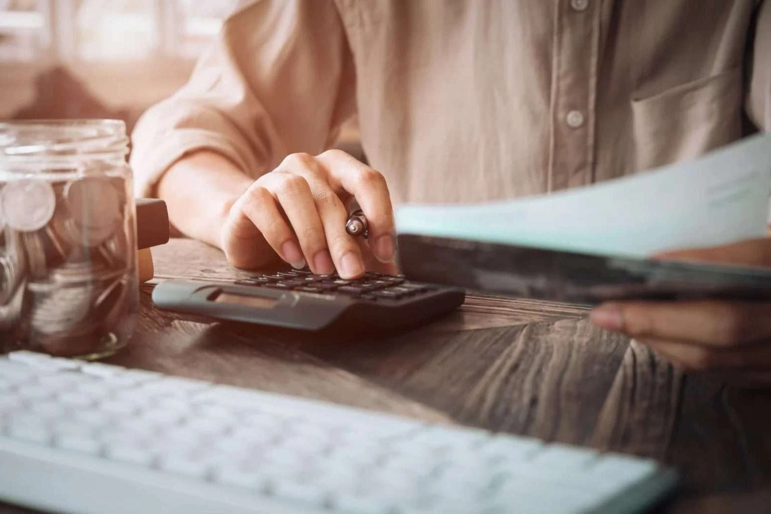 A close-up shot of a person's hands as they use a black calculator on a wooden desk. One hand holds a pen while pressing the buttons, and the other holds a document, with a jar of coins and a white computer keyboard partially visible in the frame.