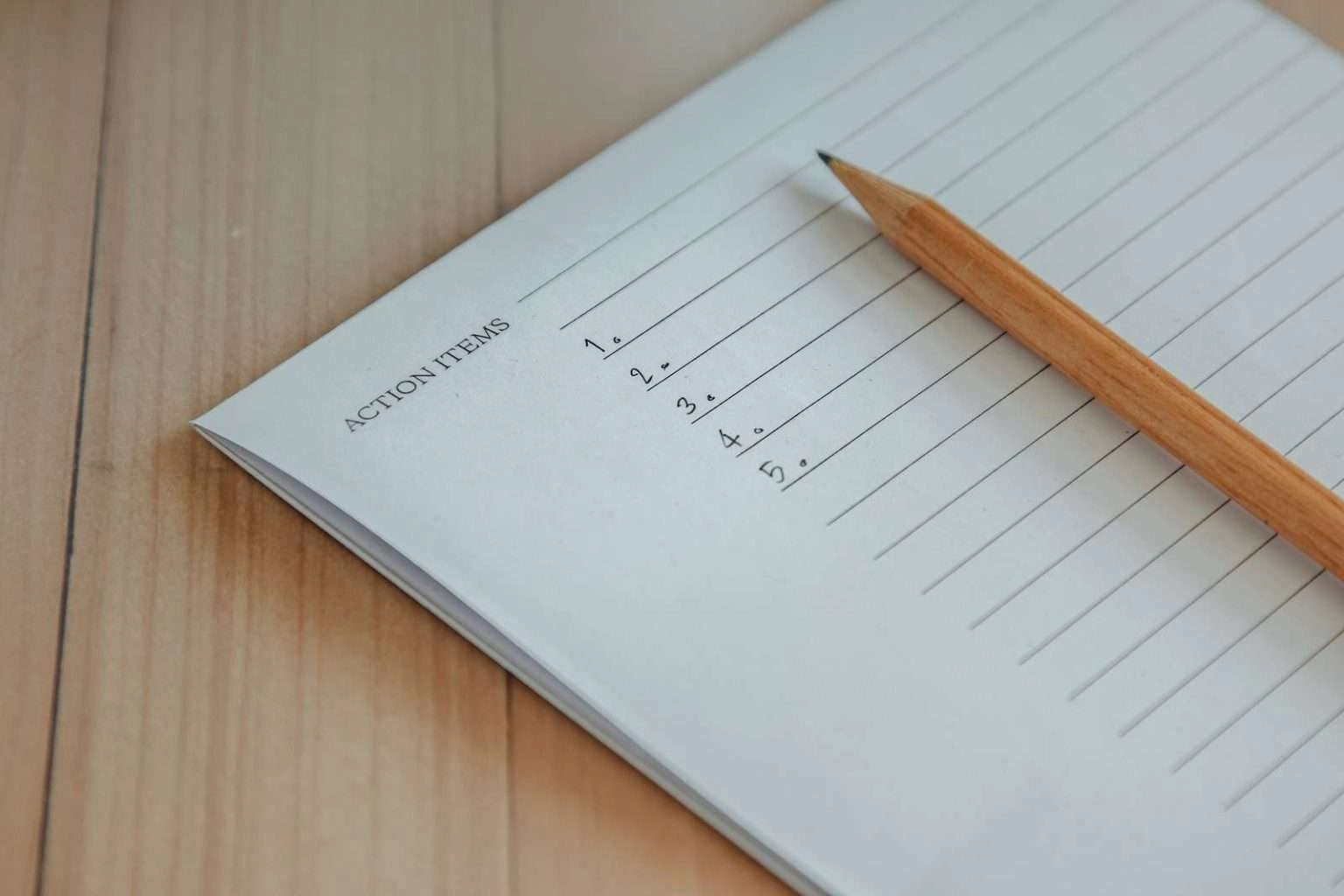 A close-up shot of a wooden pencil resting on a notepad labeled "ACTION ITEMS" with a numbered list from one to five.