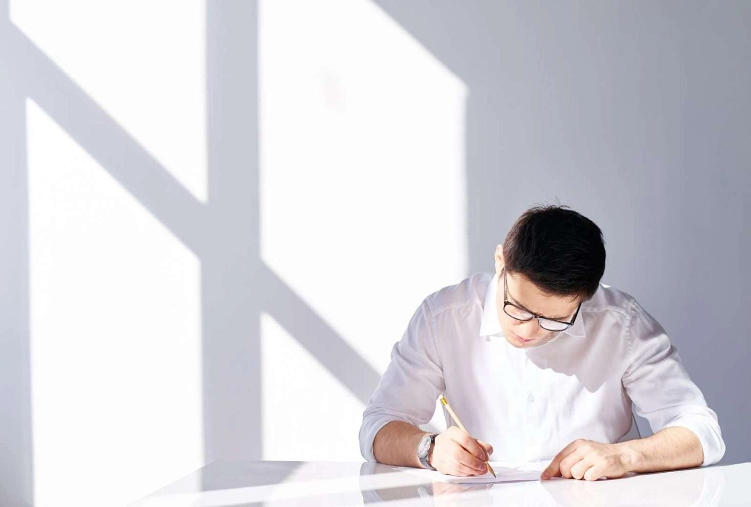 A man in a white dress shirt and glasses sits at a bright white desk, focused on writing on a piece of paper with a pencil. Sharp shadows from a window are cast on the white wall behind him.