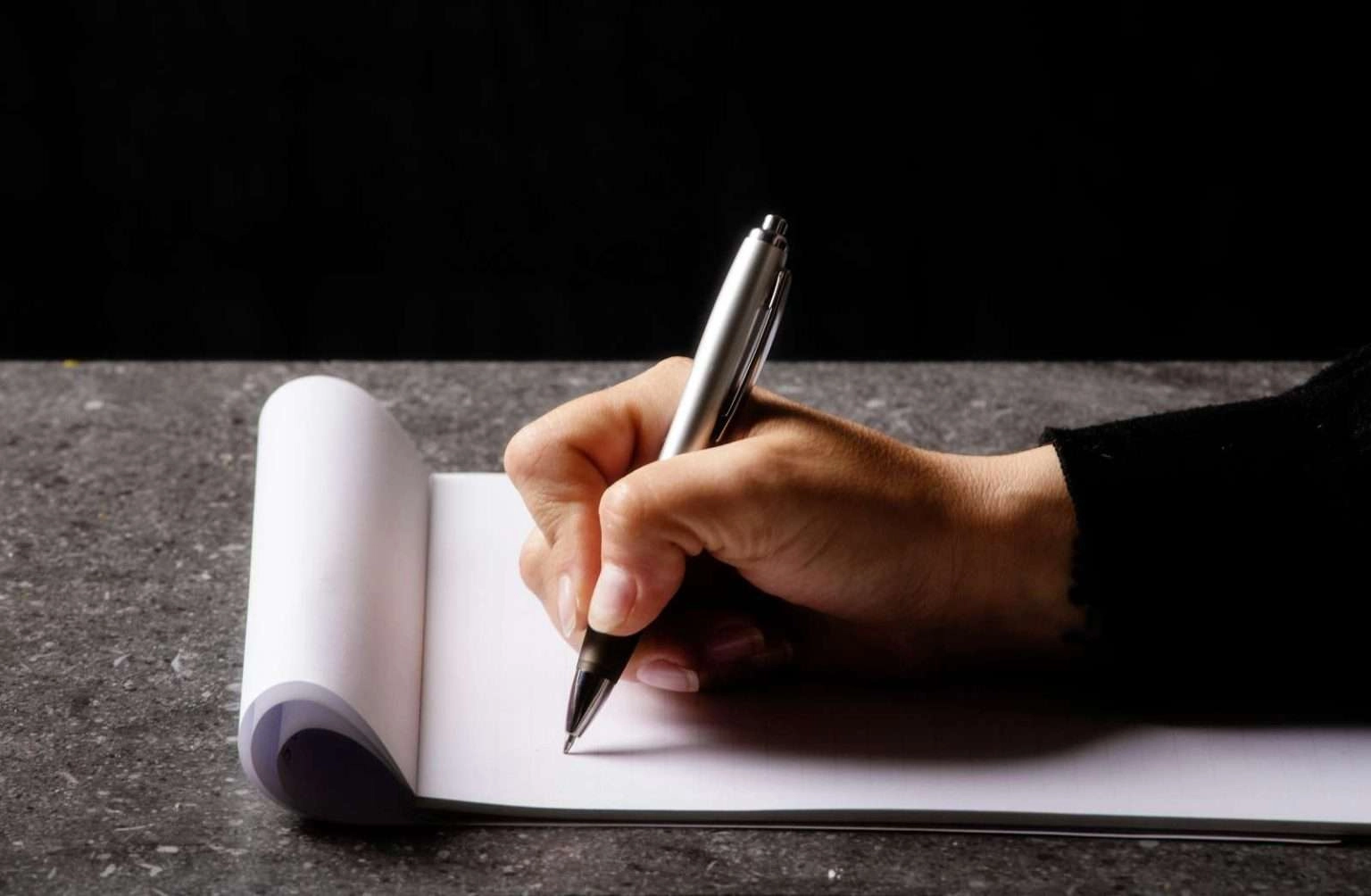 A side-profile, close-up shot of a hand holding a silver pen and writing on a white notepad with a rolled top edge, set against a dark, textured surface and black background.