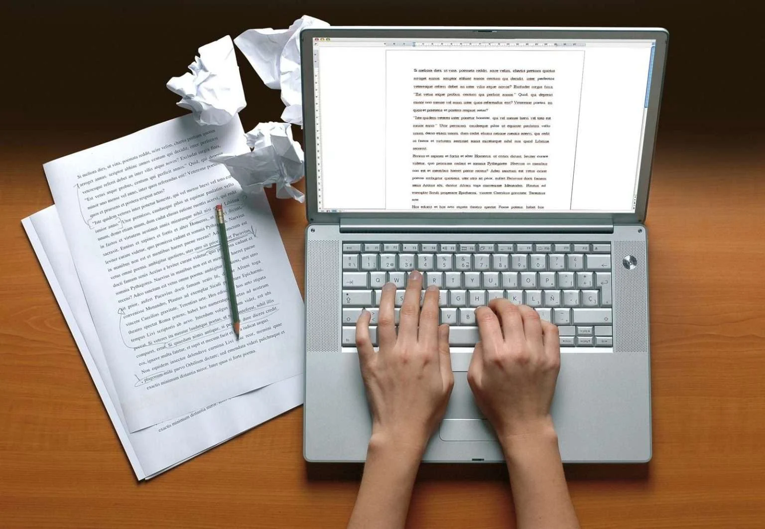 A top-down view of a person's hands typing on a silver laptop keyboard on a wooden desk, surrounded by printed pages of text and a few crumpled paper balls.
