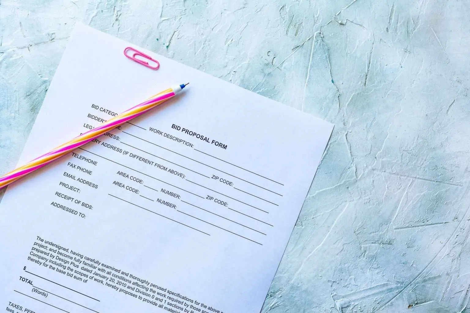 A top-down view of a blank "BID PROPOSAL FORM" on a textured light blue surface, with a pink paper clip at the top and a colorful striped pen resting on the left side.