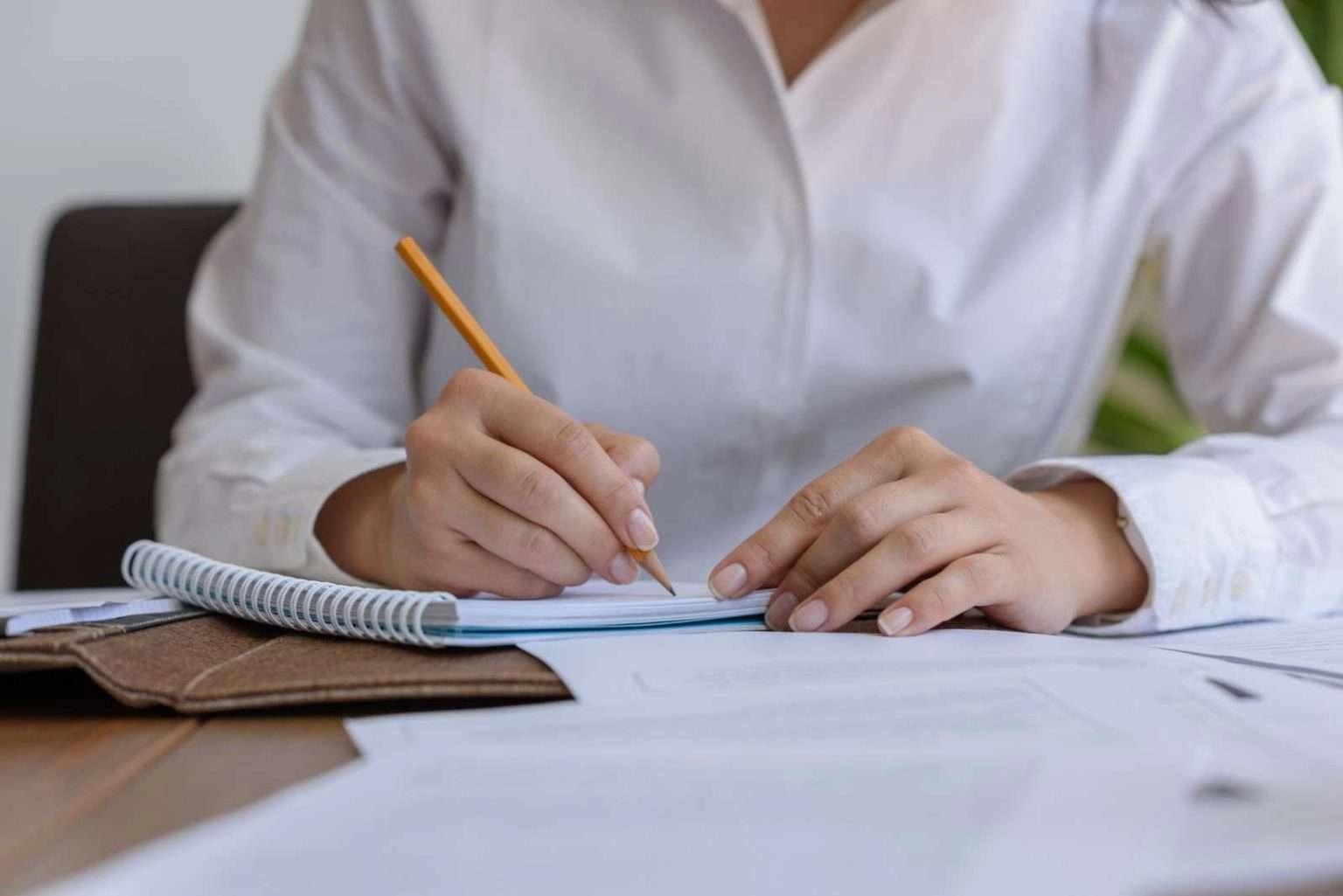 A close-up shot focuses on a person's hands as they write with a yellow pencil in a small, spiral-bound notebook resting on a brown leather-style folder.