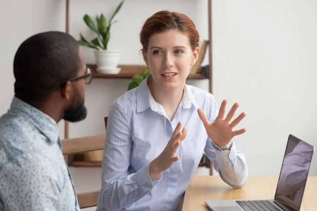 A woman with red hair wearing a light blue striped shirt sits at a wooden desk with a laptop, gesturing with her hands while talking to a man.