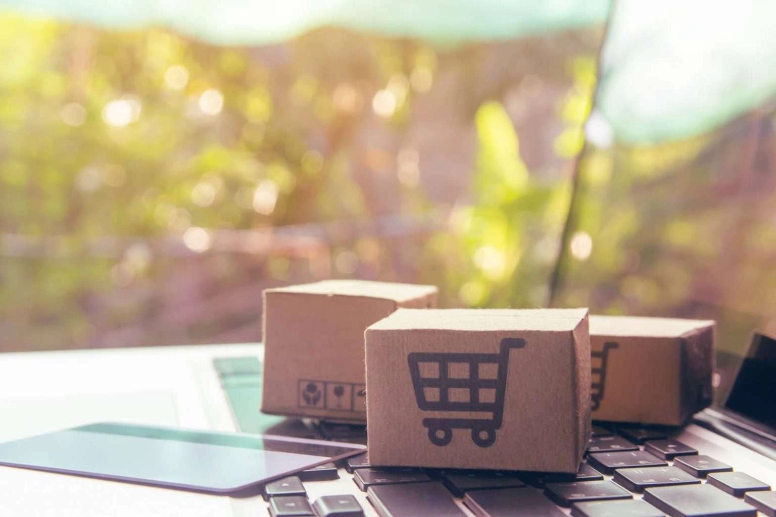 Three small cardboard boxes, one featuring a black shopping cart icon, rest on a laptop keyboard next to a credit card, with a blurred outdoor background.