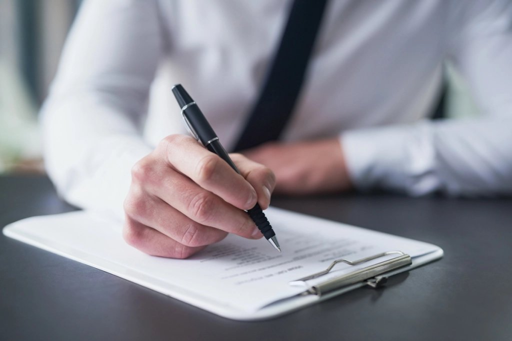 A close-up shot focuses on a person's hand in a white dress shirt and black tie, writing with a black pen on a document held in a silver clipboard.