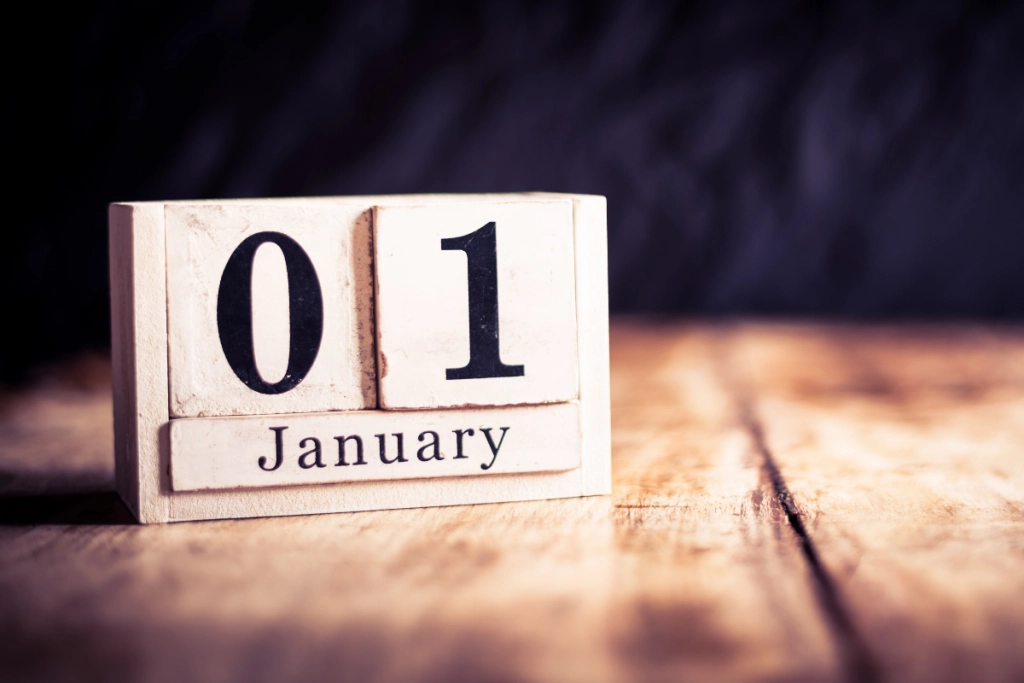 A white wooden block calendar displays "01 January" in black text, resting on a rustic wooden surface against a dark, blurred background.