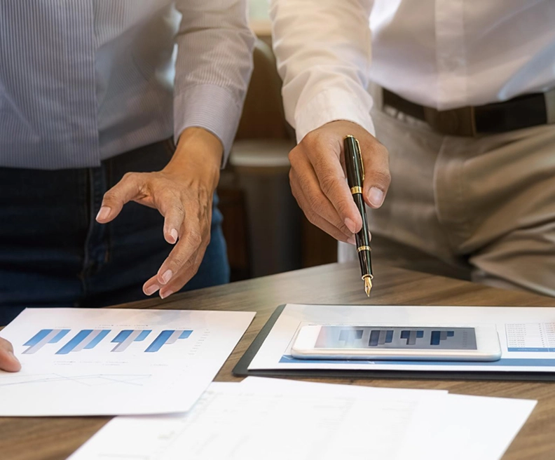 Two people stand at a wooden desk, collaborating on data analysis. One person points a gold-tipped fountain pen toward a smartphone displaying a bar chart, while the other person's hand is open near a printed document featuring similar blue and grey bar gr