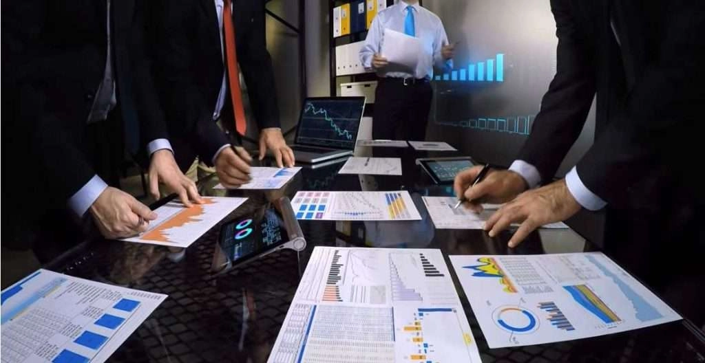 A team of professionals in business attire gathers around a glass conference table covered in various printed data reports, charts, and digital tablets. Several people are actively writing on the documents or pointing at the analytics