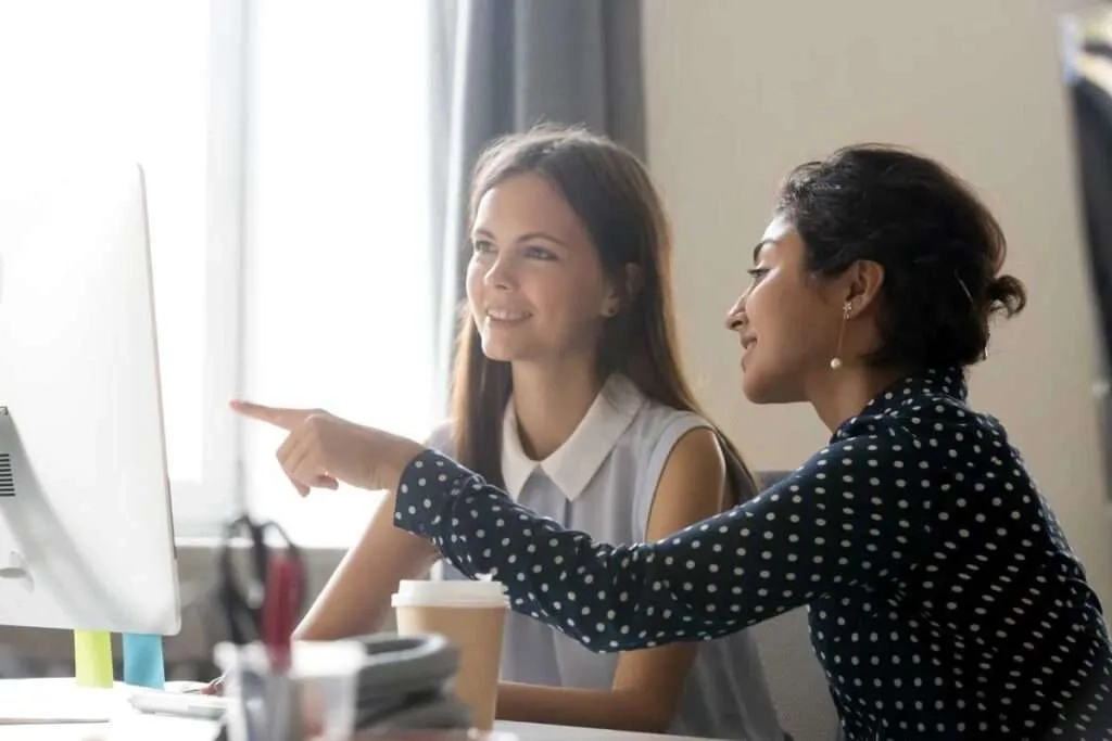Two young women are working together in a bright office; one points toward a computer screen while they both smile and look at the monitor.