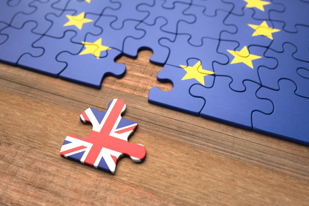 A single puzzle piece featuring the Union Jack flag lies on a wooden surface, separated from a larger blue jigsaw puzzle that forms the European Union flag with its yellow stars.