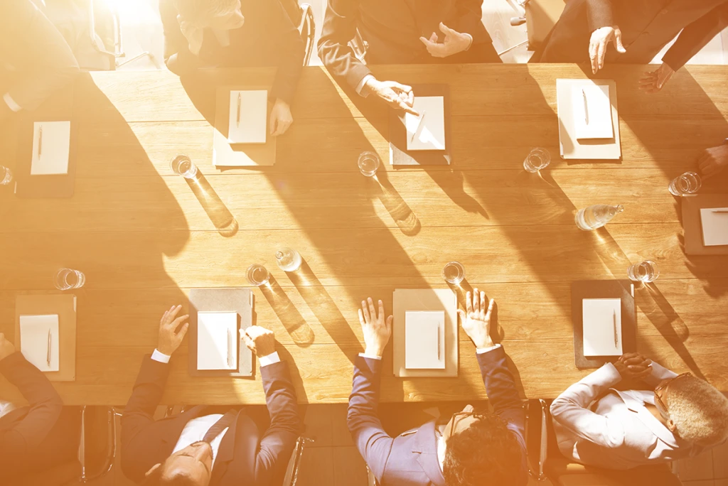 An overhead view of a diverse group of professionals in business attire seated around a long wooden boardroom table, with each person having a notepad and pen in front of them, all bathed in warm, bright sunlight.