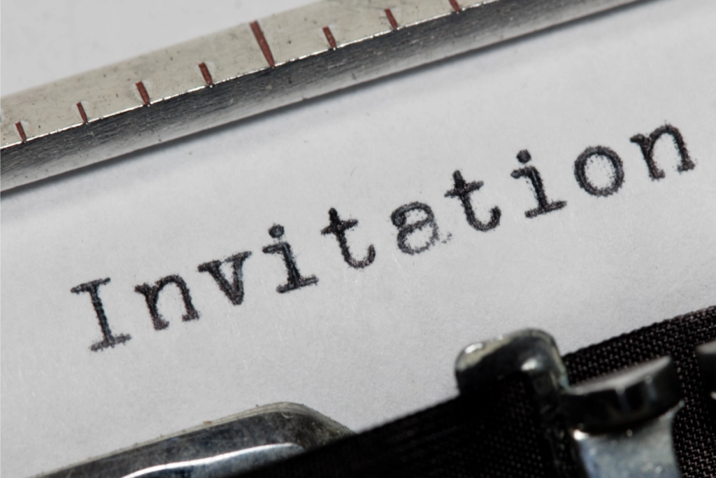 An extreme close-up of the word "Invitation" typed in a classic typewriter font on a piece of white paper, with part of the typewriter's metal carriage and ribbon visible.