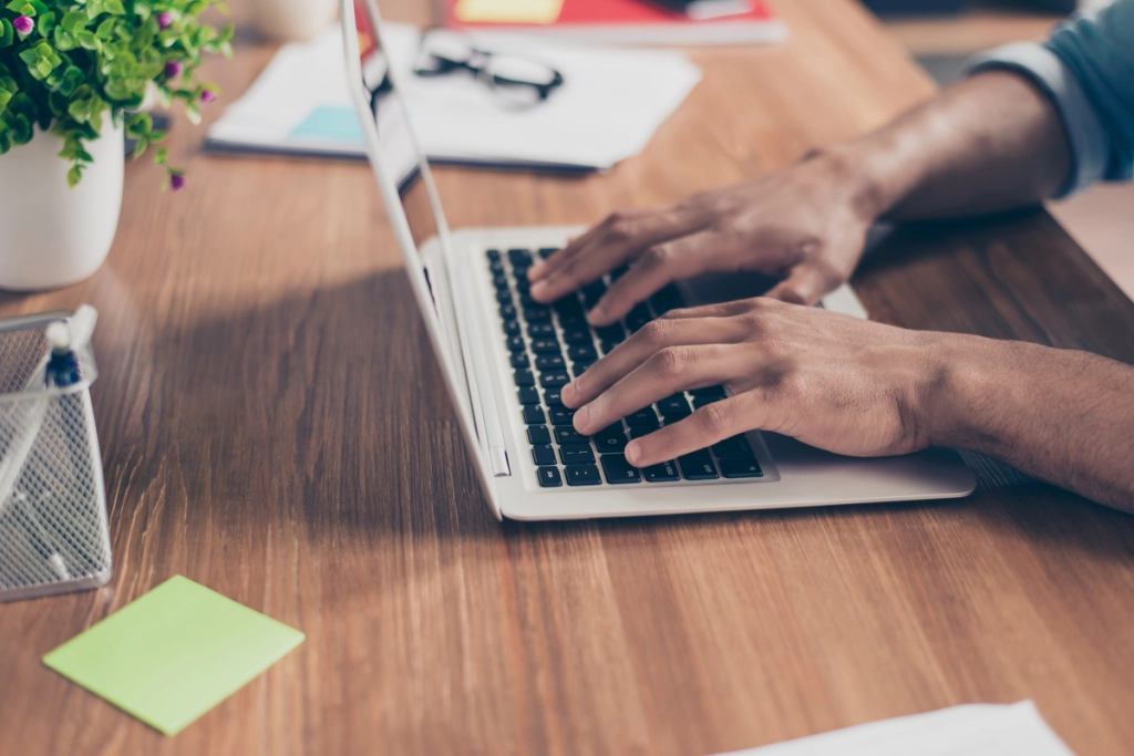 A close-up, angled view of a person’s hands typing on a silver laptop keyboard at a wooden desk, with a small potted plant and office supplies visible in the blurred background.