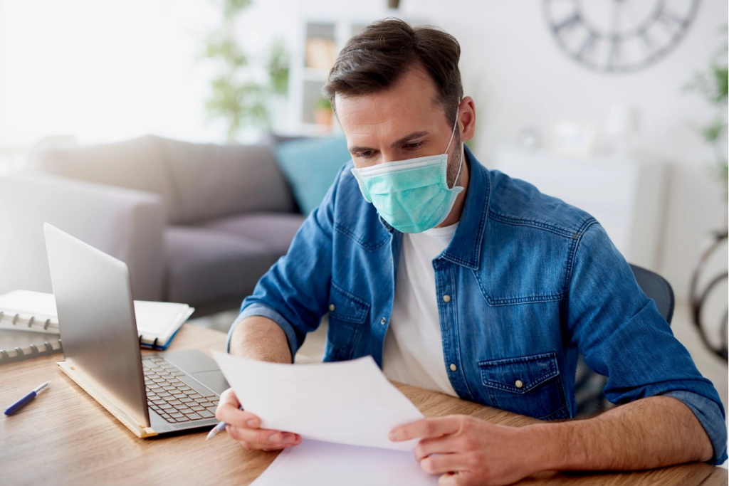 A man wearing a blue denim shirt and a light green medical face mask sits at a wooden desk, holding and reviewing a white document while working on a laptop in a bright home office setting.