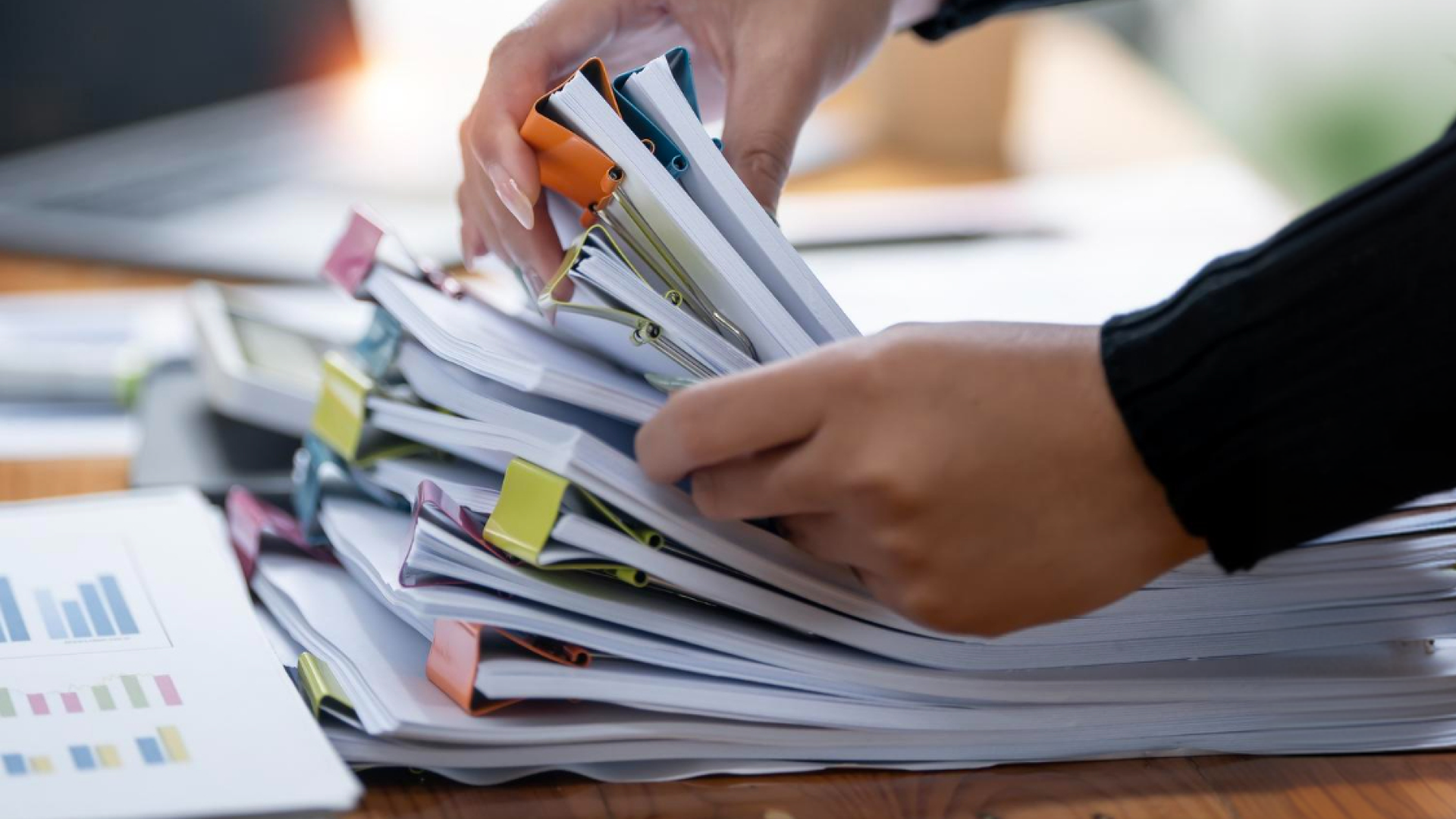 A close-up of two hands flipping through a large stack of documents secured with colorful binder clips, with a printed bar chart visible on the desk beside them.