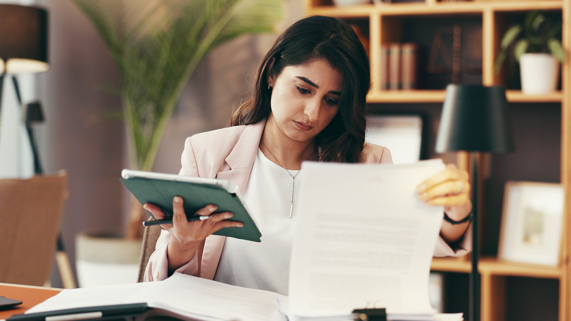 A woman in a pink blazer sits at a desk reviewing a printed document in one hand and holding a tablet in the other, with a bookshelf, desk lamp, and potted plant visible in the background.