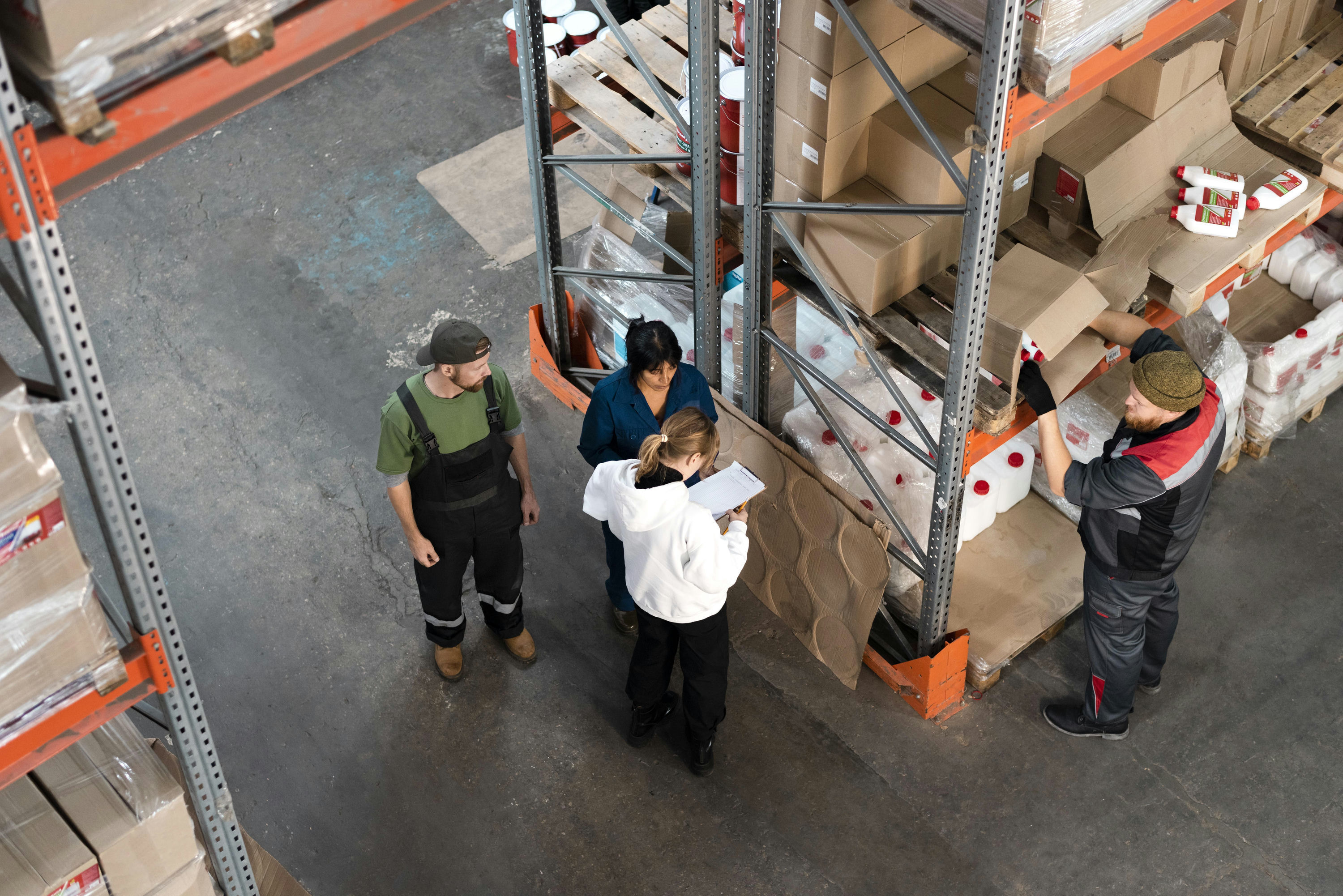 An overhead view of four warehouse workers standing between tall metal shelving units stacked with cardboard boxes and containers, with one worker writing on a clipboard and another pulling items from a shelf.
