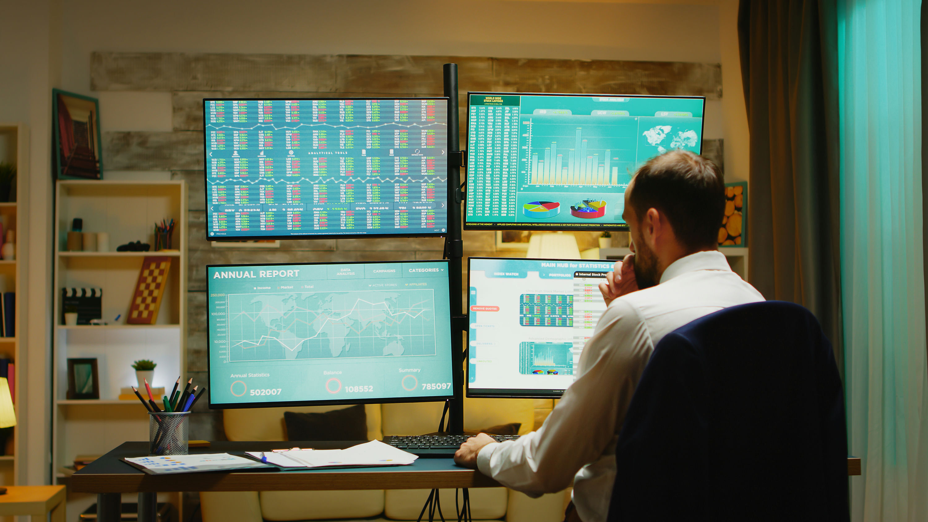 A man in a white dress shirt sits with his back to the camera, reviewing four monitors mounted on a stand displaying data dashboards, charts, a world map, and an annual report with statistics, in a warmly lit home office setting.