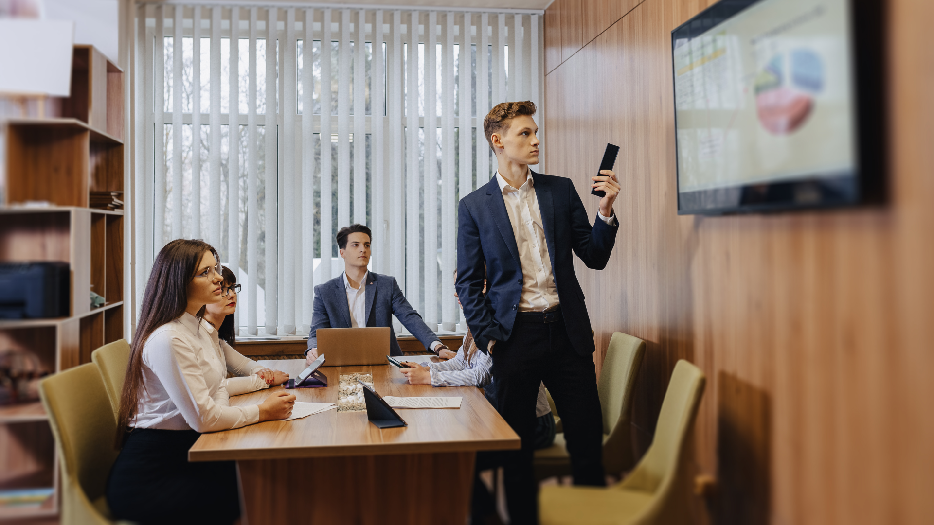 Team in a conference room watching a colleague present data on a wall-mounted screen.