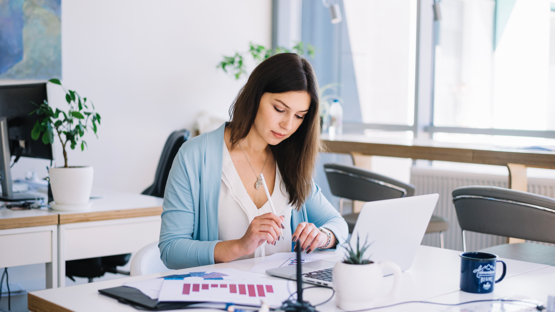 Professional working at a desk with a laptop, printed charts, and a coffee mug.