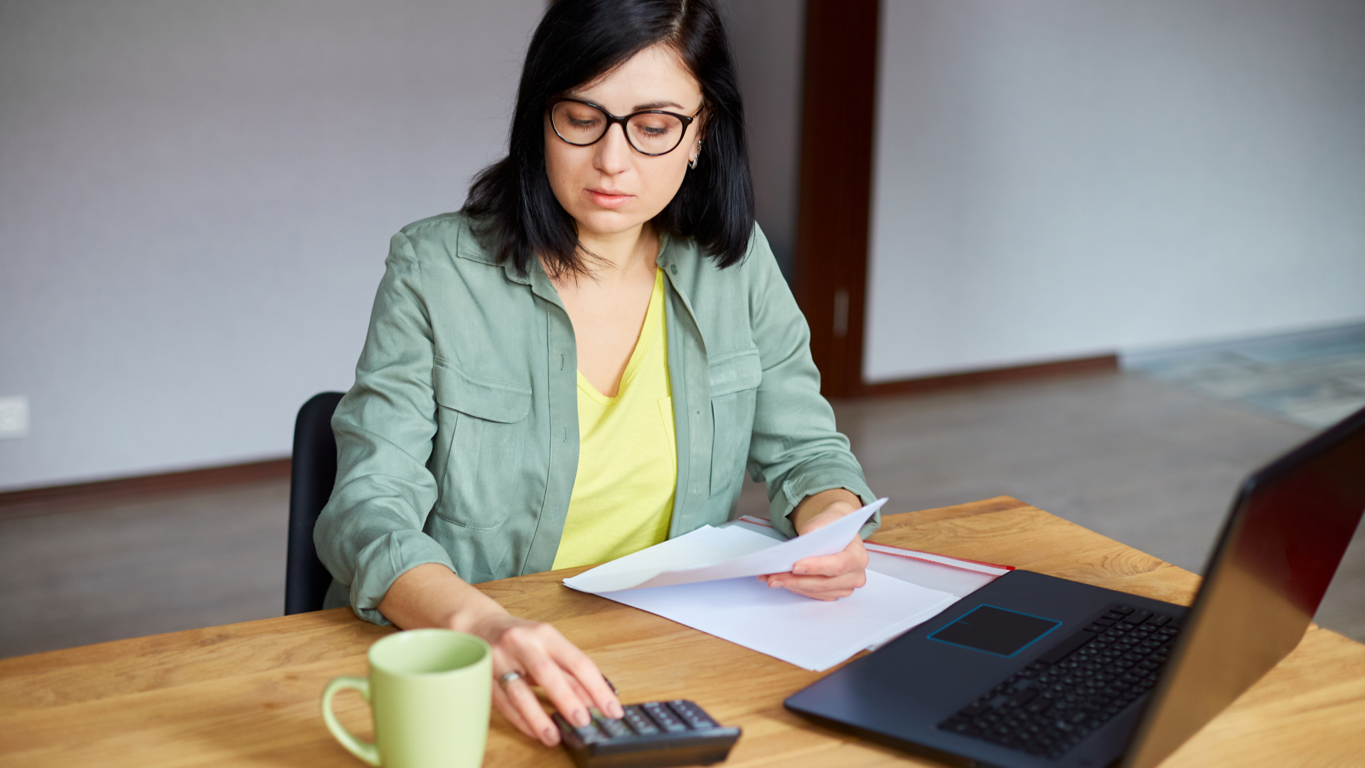 Professional reviewing documents and using a calculator at a desk with a laptop and coffee mug.