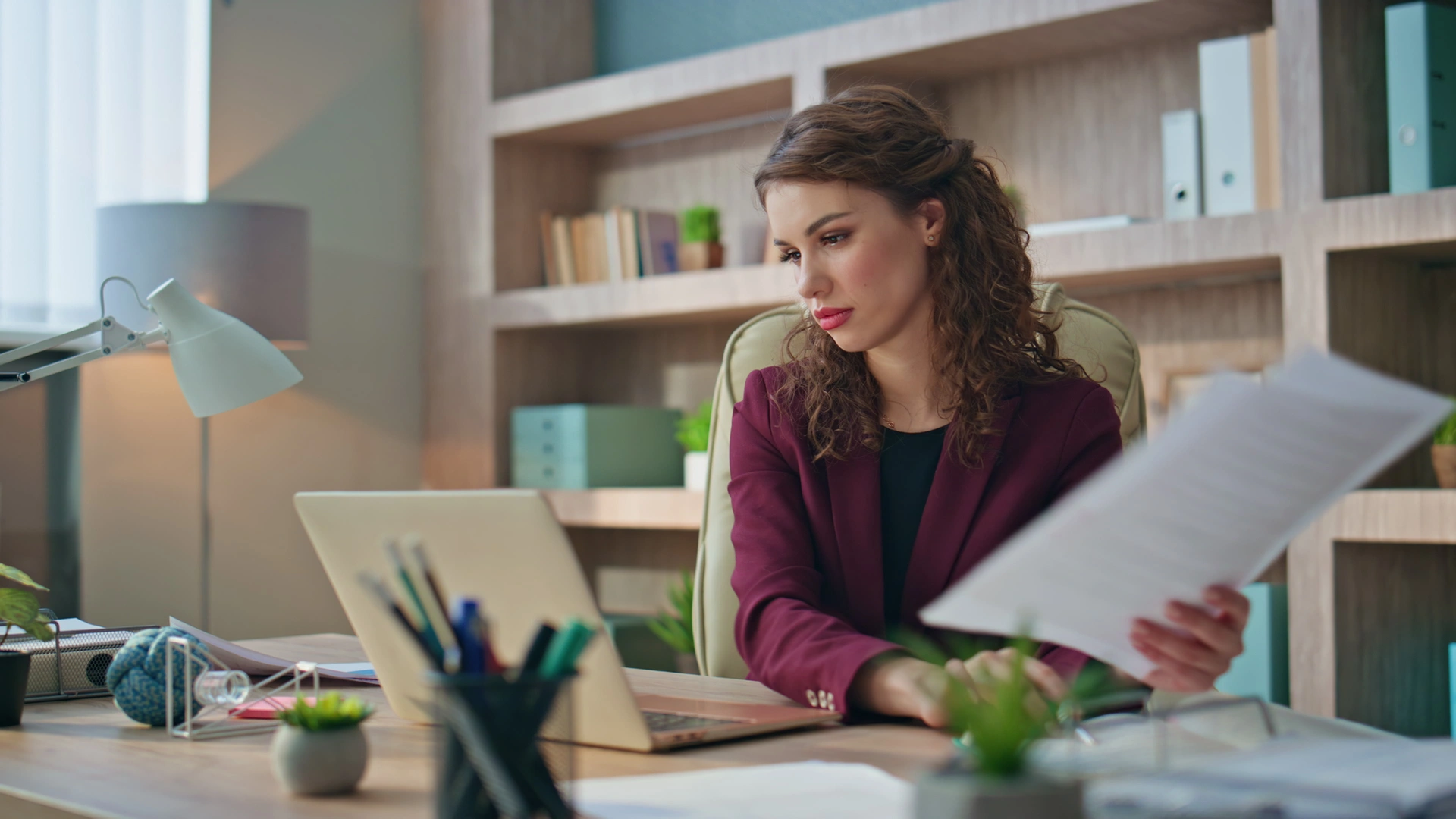 A businesswoman in a maroon blazer reviews papers and a laptop at her desk.