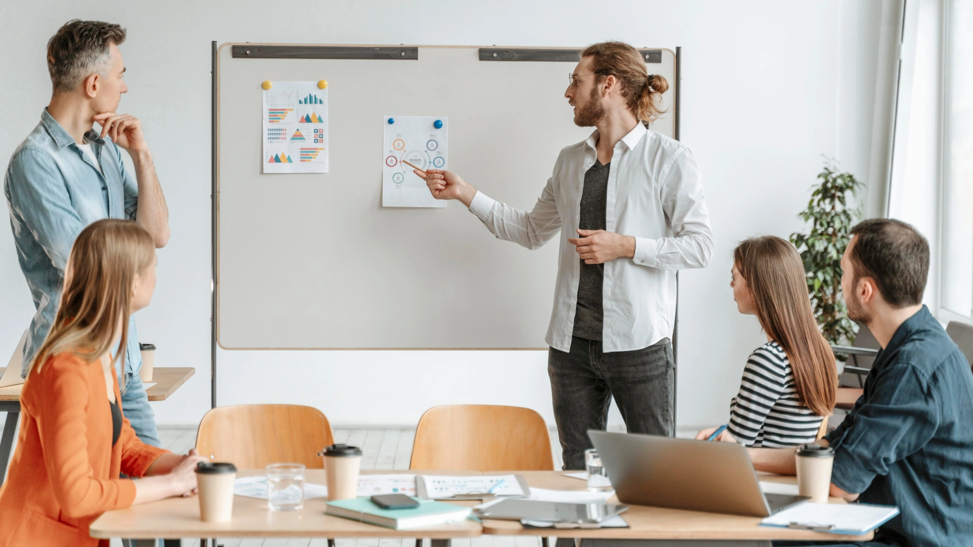 A professional stands at a whiteboard pointing at a chart. Four colleagues pay attention: one standing, and the three others sitting