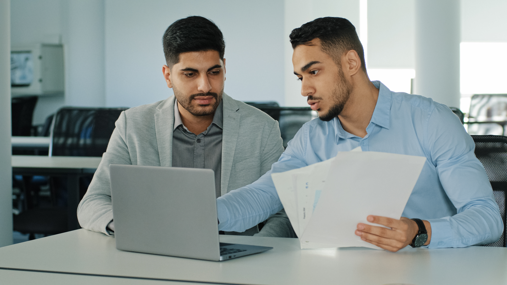 Two colleagues reviewing a laptop and printed reports to weigh deduction management software criteria.