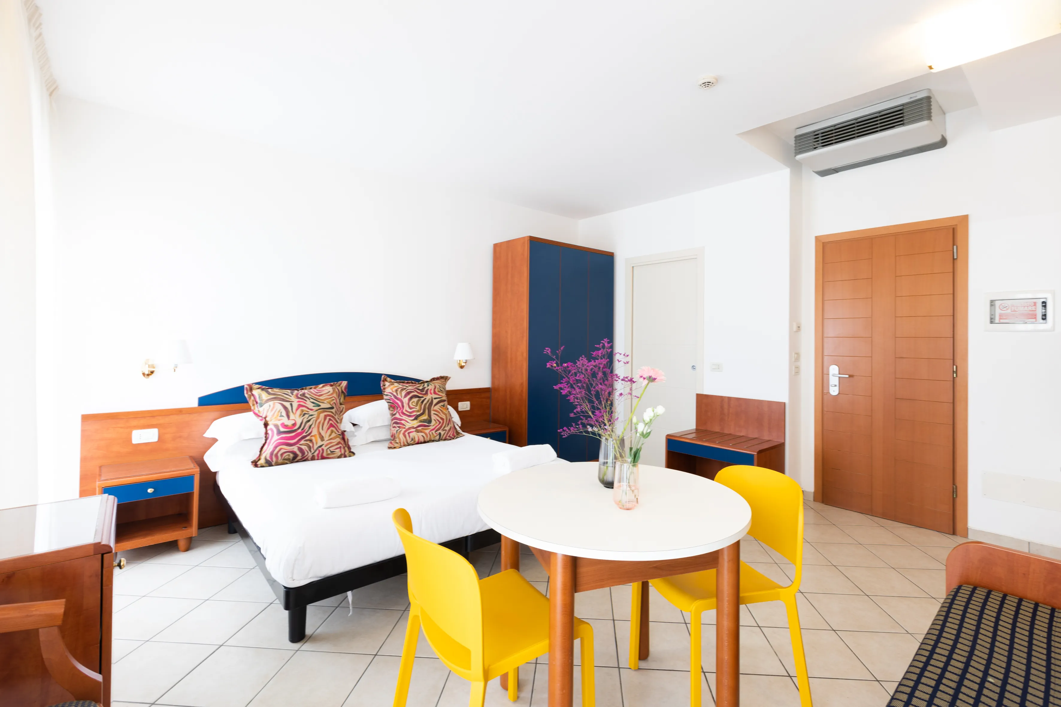 Camera d'hotel luminosa con letto matrimoniale, cuscini decorativi, tavolo rotondo con due sedie gialle e fiori, armadio e aria condizionata.