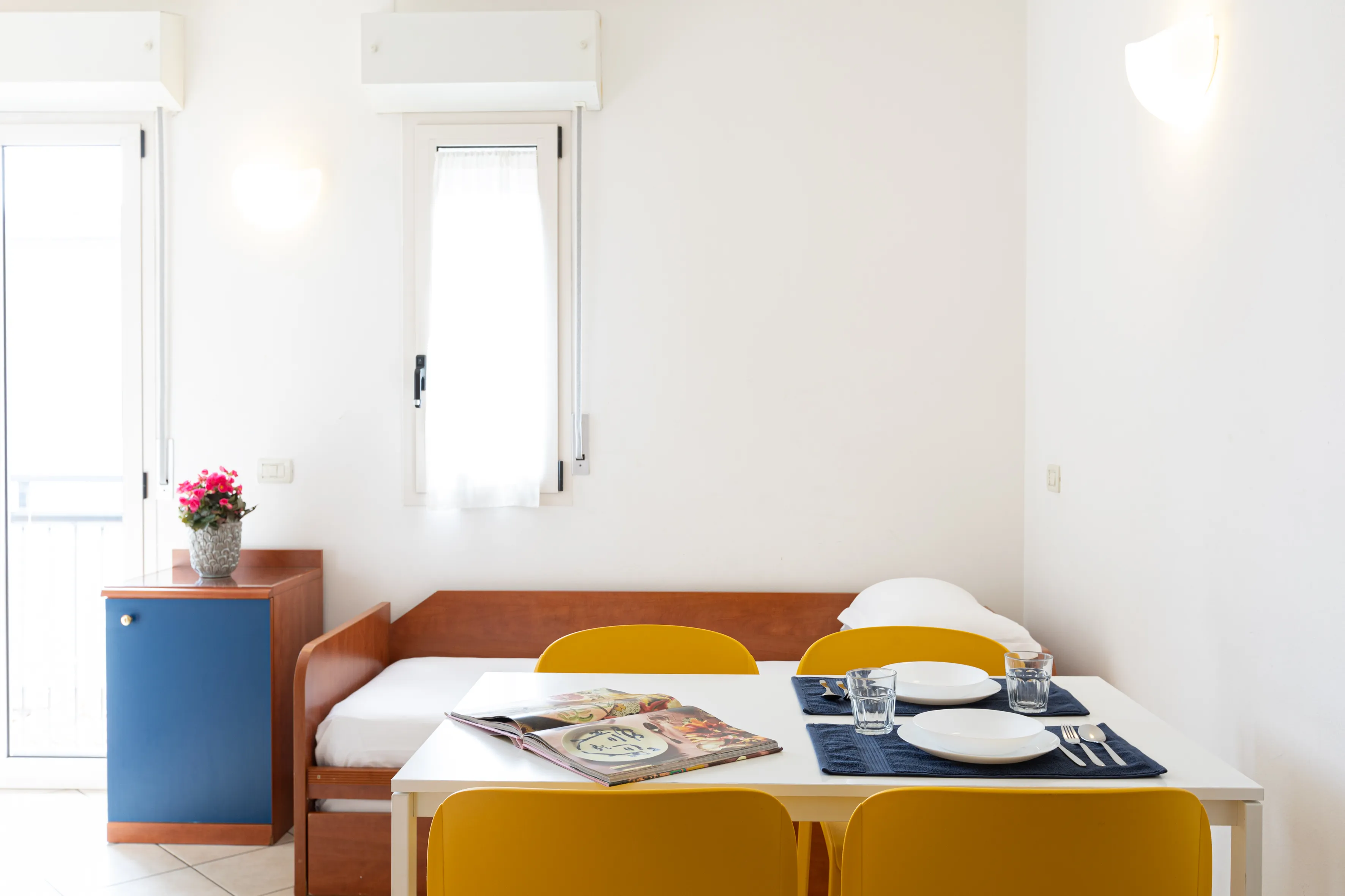 Soggiorno luminoso con tavolo bianco apparecchiato per due persone e sedie gialle, vicino a divano letto in legno e mobiletto con vaso di fiori.